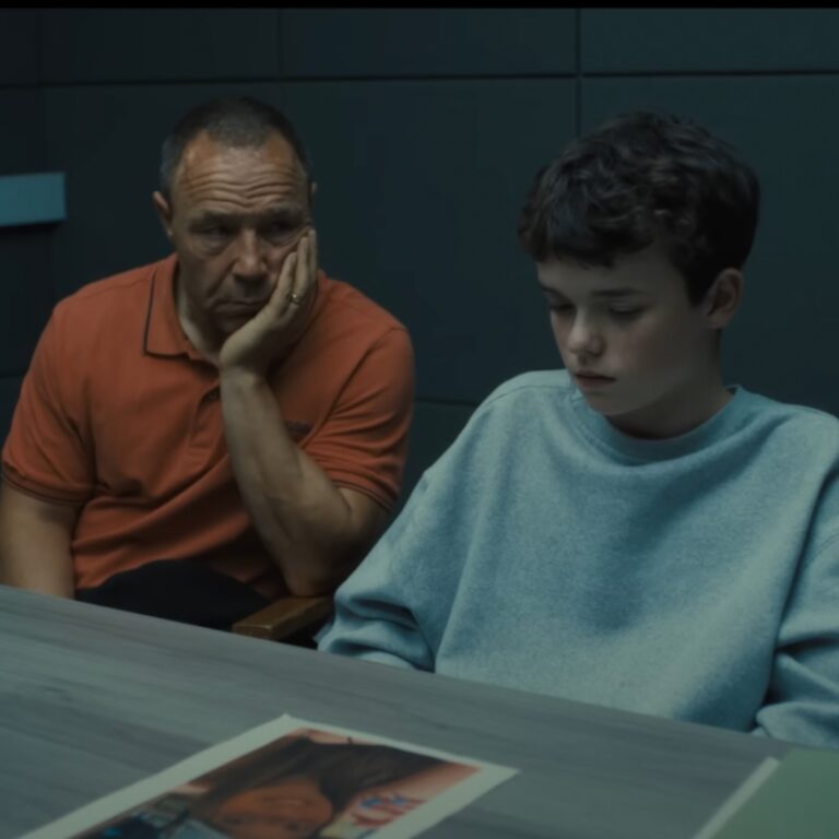 Scen uit de Netflix-serie Adolescence: een man zit naast een tienerjongen in een kamer bij een tafel. De man heeft zijn hand onder zijn kin en kijkt naar de jongen. De jongen kijkt naar een foto op de tafel van een meisje die lachend naar de camera kijkt.