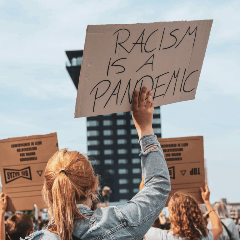 Persoon met een paardenstaart houdt een bord omhoog tussen een groep mensen die ook kartonnen borden omhooghouden. De tekst op het bord is: 'racism is a pandemic'.