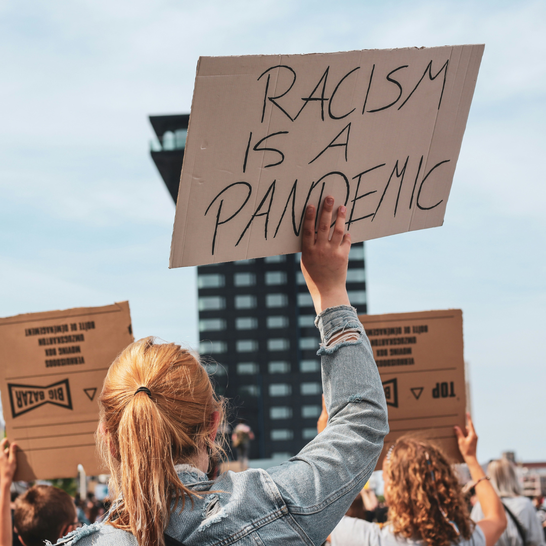 Persoon met een paardenstaart houdt een bord omhoog tussen een groep mensen die ook kartonnen borden omhooghouden. De tekst op het bord is: 'racism is a pandemic'.