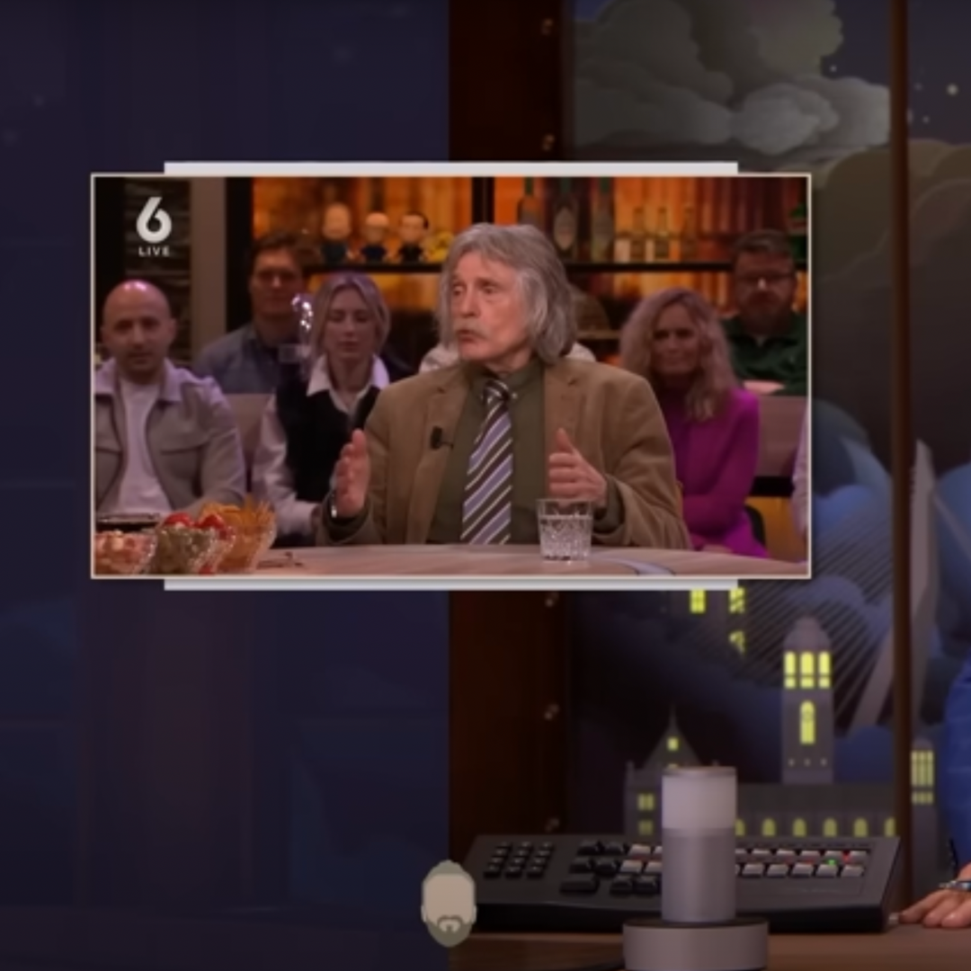 Screenshot van LUBACH. Arjen Lubach zit in de studio en heeft zijn hand voor zijn gezicht, naast hem is een klein scherm te zien met daarop Johan Derksen.