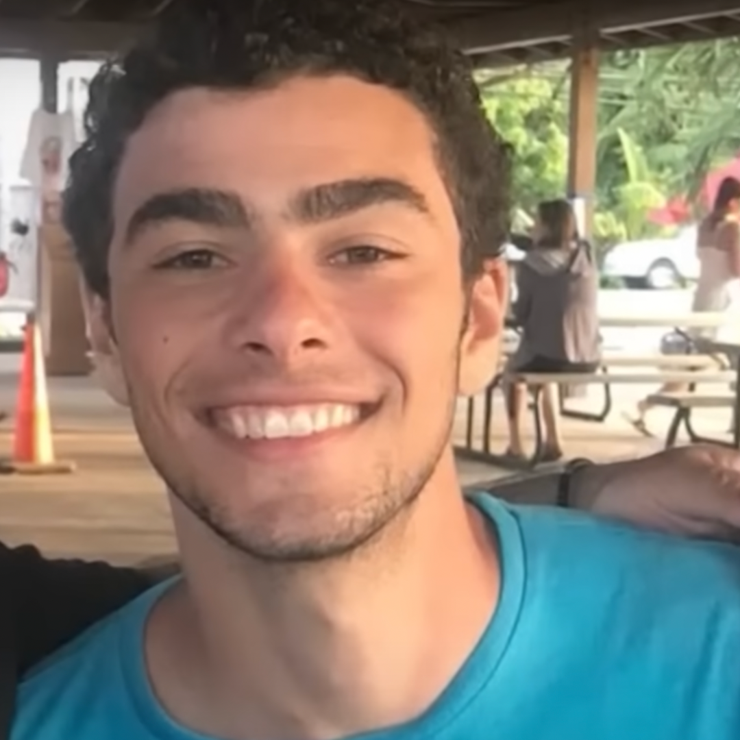 Luigi Mangione kijkt lachend naar de camera met een hand om zijn schouder. Op de achtergrond zijn mensen en picnic tafels te zien.