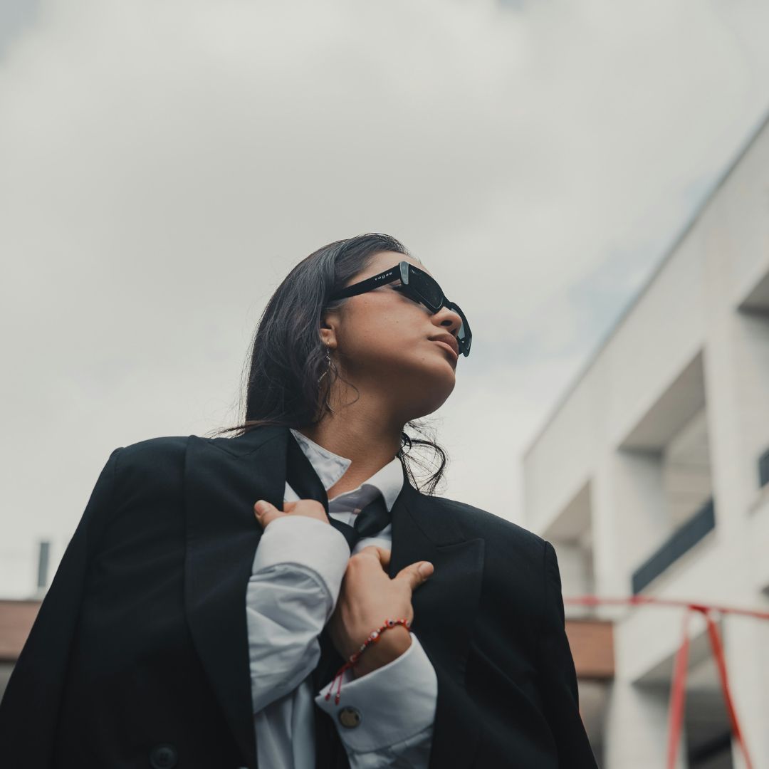Een persoon met zwarte blazer en zwarte zonnebril draagt een zwarte stropdas losjes om de nek. De foto is gemaakt van beneden en de lucht achter is grijs