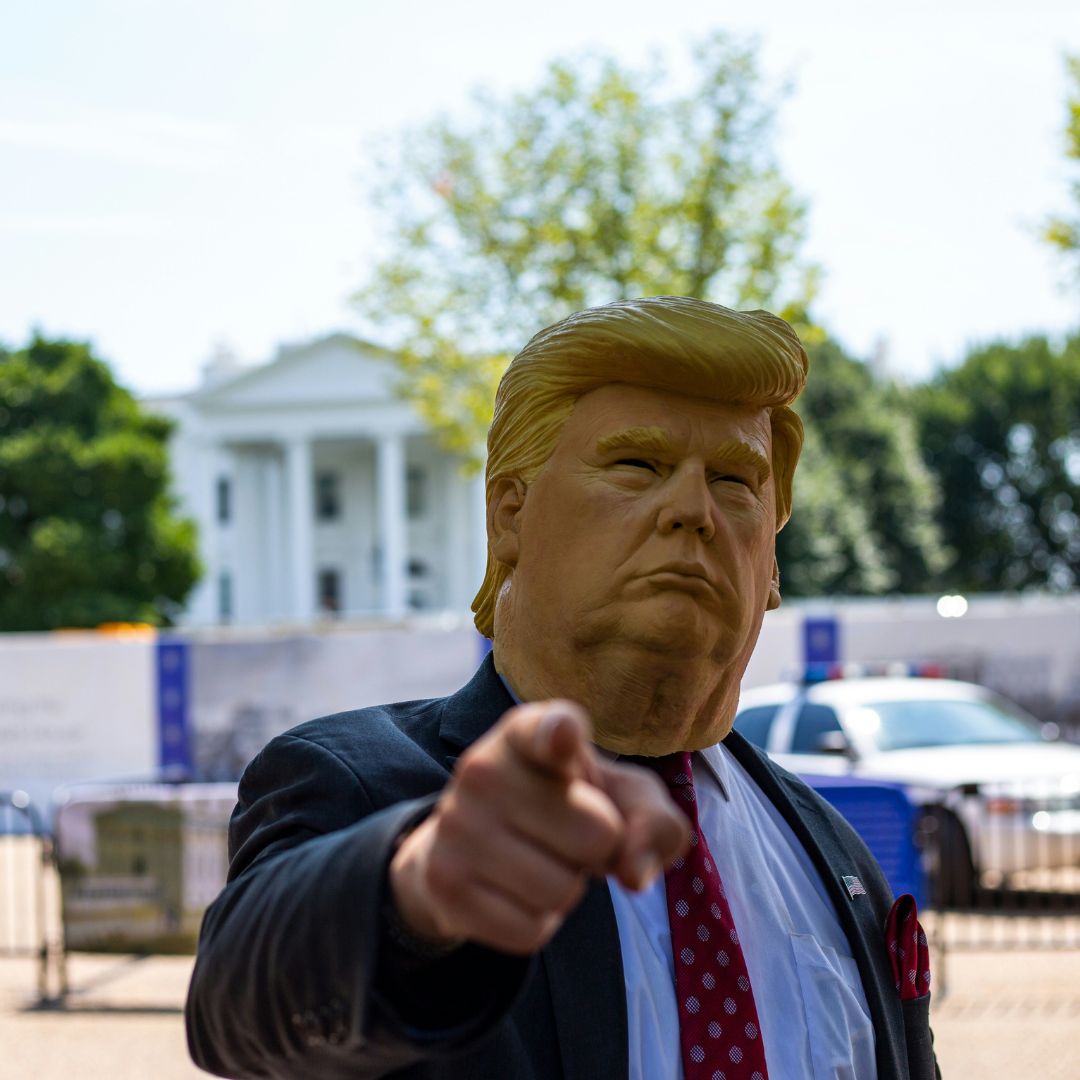 Iemand met een realistisch Trump masker op staat voor het Witte Huis, in pak, en wijst naar de camerav