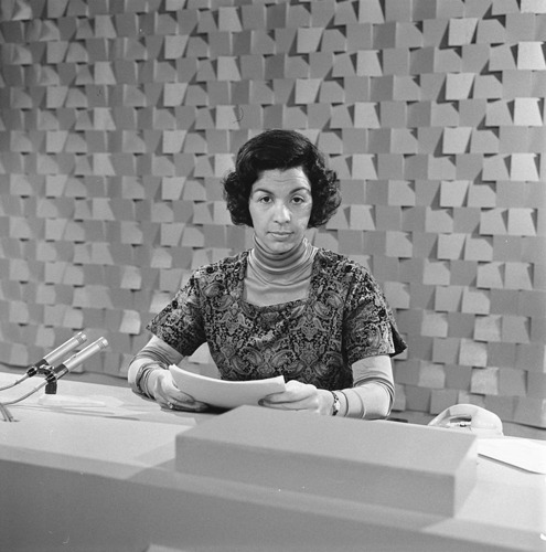 Zwart-wit foto van Eugène Herlaar achter haar bureau en microfoon.